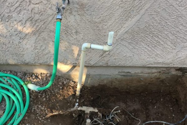 Outdoor irrigation system with exposed pipes, valves, and a green hose next to a dug-up section of soil by a wall.