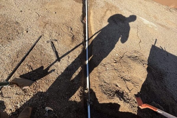 A person’s shadow falls over a narrow trench with exposed piping and excavation tools on the dirt ground.