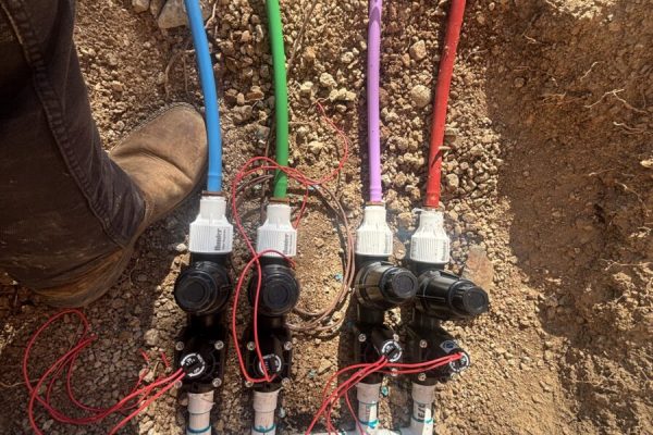 Three irrigation valves connected to color-coded wires and PVC pipes are partially buried in dirt, with workers’ boots visible.