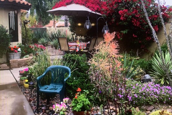 A patio with a table and chairs under an umbrella surrounded by colorful flowers, plants, and a green plastic chair.