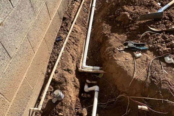 PVC pipes installed in a trench alongside a brick wall, with plumbing tools and soil surrounding the area.