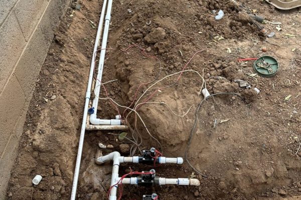 Exposed irrigation system with PVC pipes and valves installed in a trench next to a block wall on bare soil.