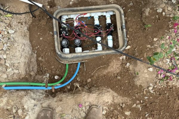 Person standing over an open irrigation valve box with exposed pipes and wiring, surrounded by dirt and rocks.