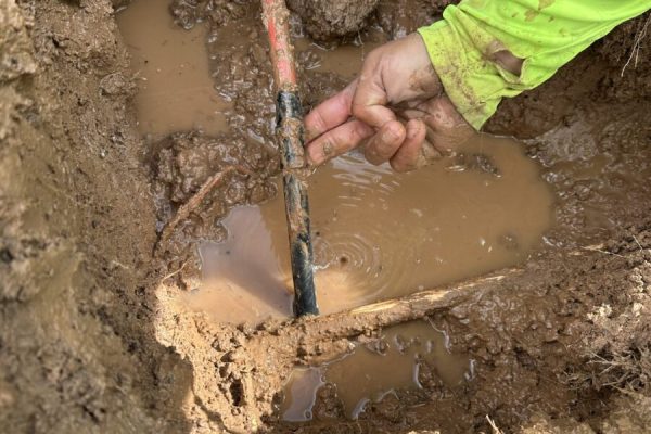 A person in a green sleeve repairs a leaking pipe in a muddy, water-filled hole in the ground.