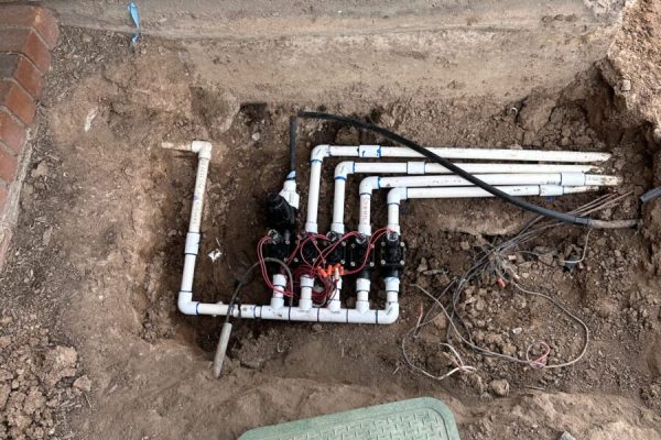 Irrigation control valves and PVC pipes are exposed in a dug-out section of ground near a brick wall and green cover.