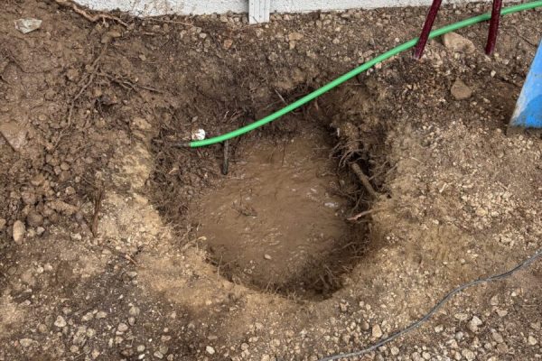 A muddy hole in the ground near a white wall, with cables, a hose, and red-handled pliers on the dirt.