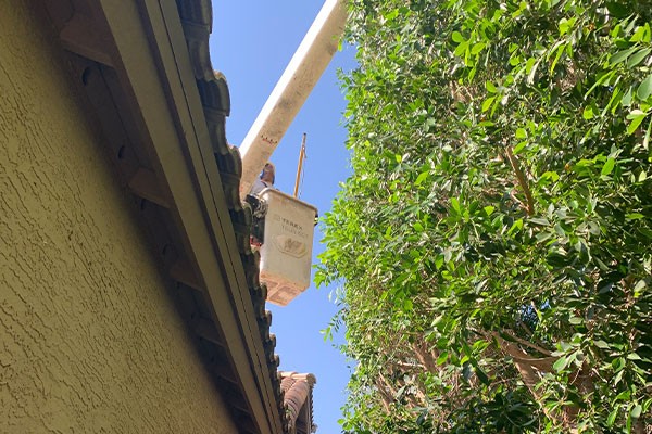 Worker in a lift bucket trims tree branches near the edge of a house roof on a sunny day.