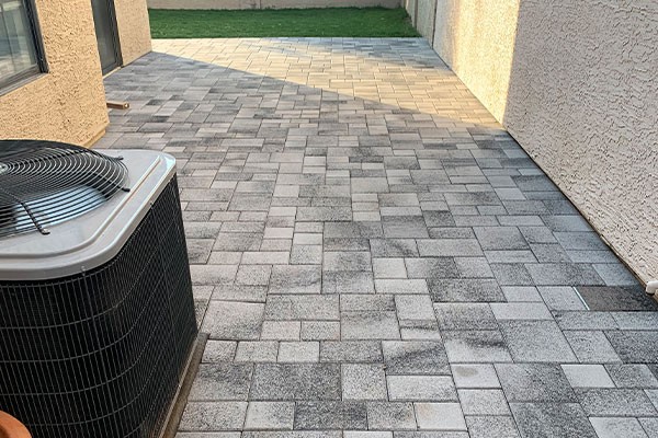Backyard patio with gray interlocking pavers, adjacent to a house wall and an air conditioning unit, with grass in the distance.