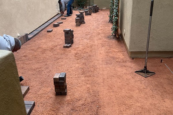 Workers are preparing a dirt pathway and stacking pavers alongside a beige building for installation.