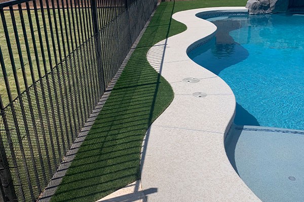 Curved poolside edge with artificial grass strip and black metal fence casting shadows on the grass.