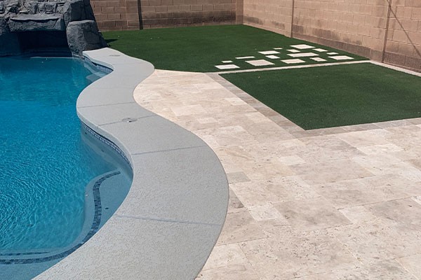 Curved poolside with stone tiles, artificial grass, and square concrete pavers in a backyard.