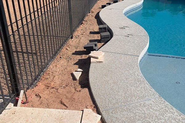 Poolside area with unfinished paving, scattered tiles, and loose bricks along a curved fence near the water.