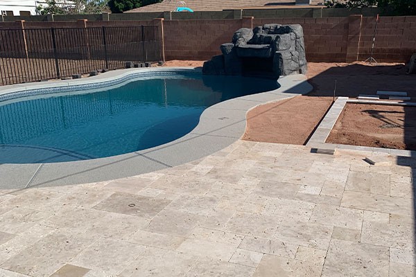 A backyard pool with a stone waterfall feature, partially paved patio, and unfinished landscaping with dirt areas.