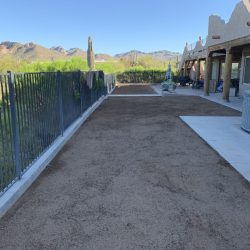 A fenced yard with a gravel path, concrete walkway, and desert landscape next to a stucco building under a clear sky.