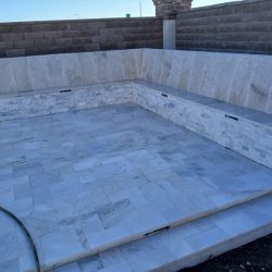 Empty, unfinished pool with light-colored stone tile and a garden hose lying on the floor, surrounded by stone walls.