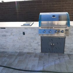 Outdoor kitchen with a built-in grill, sink, and stone countertop against a brick wall on a tiled patio.