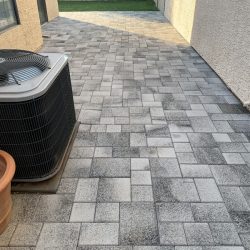 A paved patio with gray stone tiles next to a beige building, an air conditioning unit, and a small patch of grass in the distance.