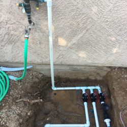 Outdoor irrigation valve system with PVC pipes and hoses attached, partially buried near a stucco wall.