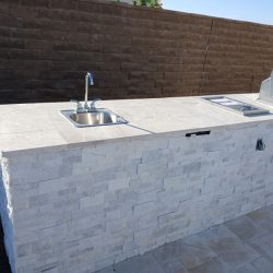 Outdoor kitchen with a stone countertop, built-in sink, and grill against a brown brick wall.