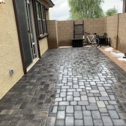 A wet stone patio next to a house with a bicycle, planter, and firewood stacked against a tan brick wall.