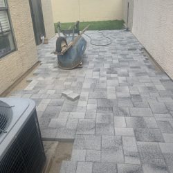 A wheelbarrow sits on a partially completed patio with gray pavers beside two beige buildings.