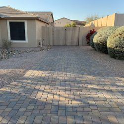 Brick-paved driveway leads to a wooden gate, bordered by gravel, bushes, and a beige house.