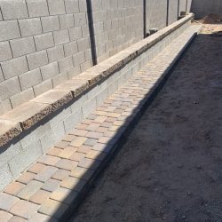 A backyard with a gray block wall, a raised brick border, and a paved walkway next to bare dirt.