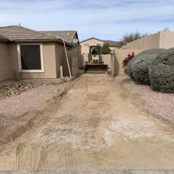A small excavator levels dirt in a cleared area between a house and a privacy wall in a residential backyard.