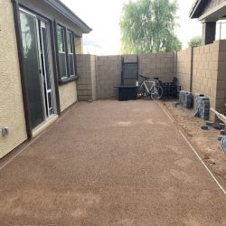 A backyard patio area with freshly leveled gravel, stacked pavers, a wheelbarrow, bike, and storage bin against a wall.