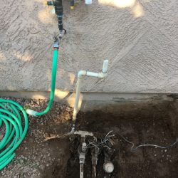 Outdoor irrigation system with exposed pipes, valves, and a green hose next to a stucco wall.
