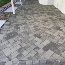 Gray herringbone-pattern pavers form a patio walkway next to a white building with landscaping on the left.