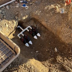 Four irrigation valves are installed in a rectangular pit in dry soil, with tools and parts nearby.