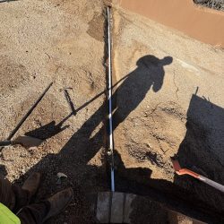 A person’s shadow falls over a partially buried pipe in a dirt trench, surrounded by tools and construction materials.