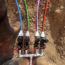 Three irrigation valves with colored wires and pipes are installed in a trench, with a worker's boots visible nearby.