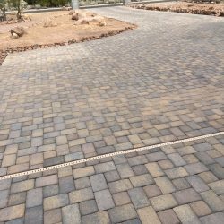 A wide driveway made of interlocking stone pavers with a drainage grate near the center, bordered by desert landscaping.