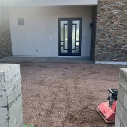 A modern house entrance with double glass doors, stone walls, and a gravel yard under construction with equipment visible.