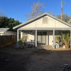 A small backyard with a wooden fence, patio furniture, potted plants, and a house with string lights on the porch.