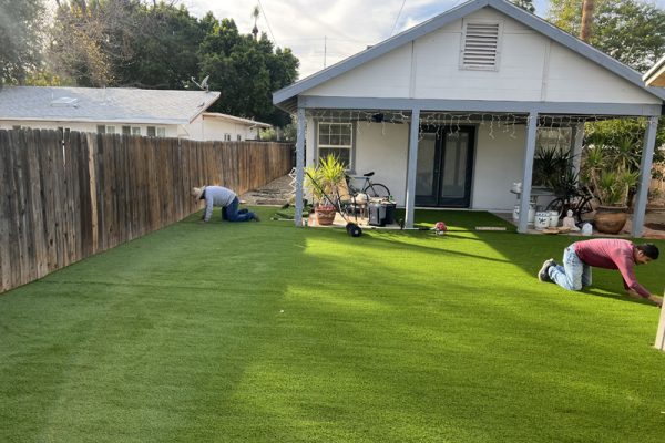 Two people install artificial grass in the backyard of a house with a covered porch and potted plants.