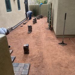 Workers are laying bricks on a red dirt ground between two beige buildings, with stacks of bricks along the sides.
