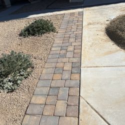 A stone paver walkway runs between gravel landscaping and a concrete driveway in front of a house.