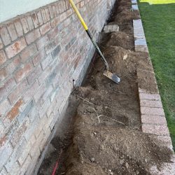 Shovel leaning against a brick wall, soil dug up along the foundation, with a red cable visible in the trench.