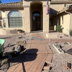 A brick walkway with missing and displaced bricks leads to the entrance of a tan stucco house with an American flag.