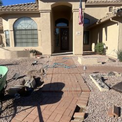 A brick walkway leading to a house is damaged, with bricks missing and tools scattered on the ground.