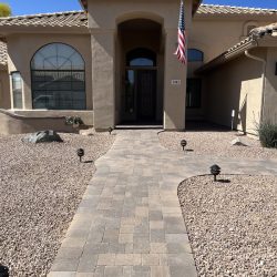 A beige stucco house with a tile roof, a brick walkway, gravel yard, and an American flag by the front door.