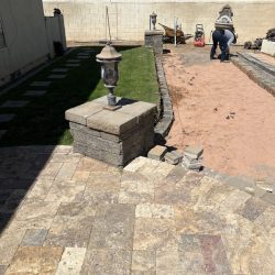 Two workers prepare a backyard area for landscaping beside a grass lawn and stone patio under clear sunlight.