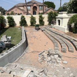 Backyard construction with a partially built block wall, arranged paver stones, and scattered construction materials.