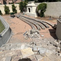 Backyard under construction with unfinished stone seating, scattered pavers, tools, and a partially built retaining wall.