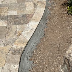 Curved stone paver edge with fresh cement border beside bare soil and small rocks in sunlight.