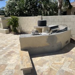 Outdoor patio with built-in grill, bench, fireplace, and tiled floor, bordered by a concrete block wall and greenery.