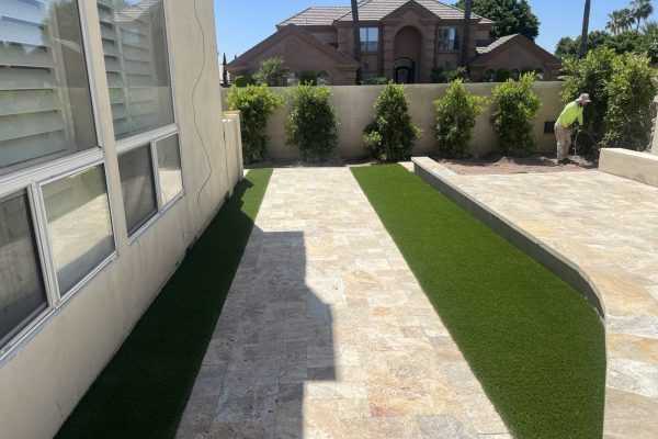 A backyard with stone tile flooring, artificial grass borders, and a person tending plants near a wall.